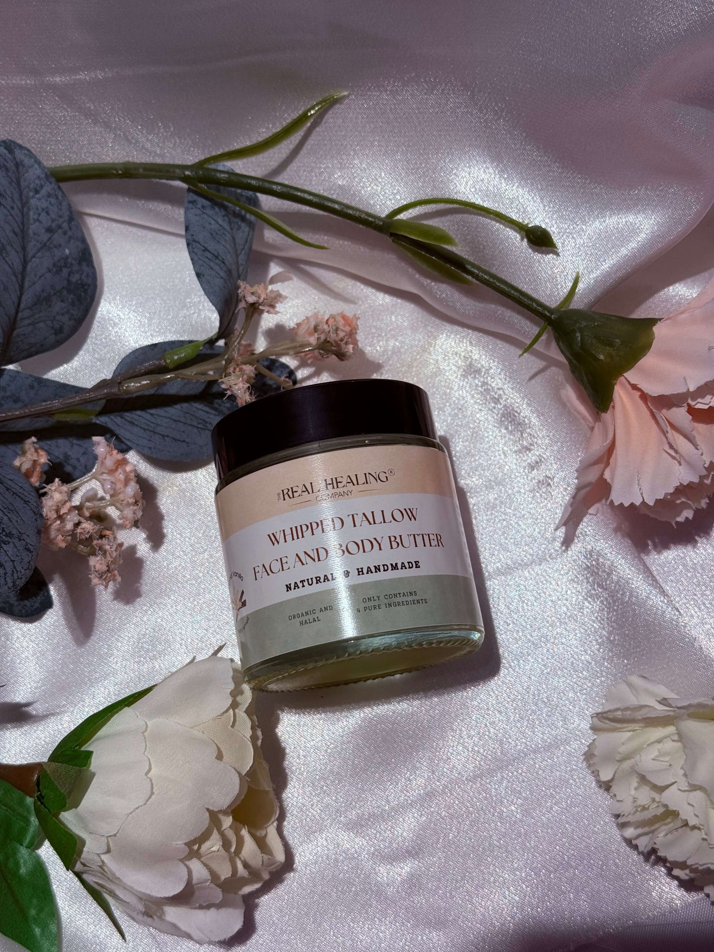 Whipped Tallow Face and Body Butter in a jar, surrounded by flowers on satin fabric, offering luxurious skin care.